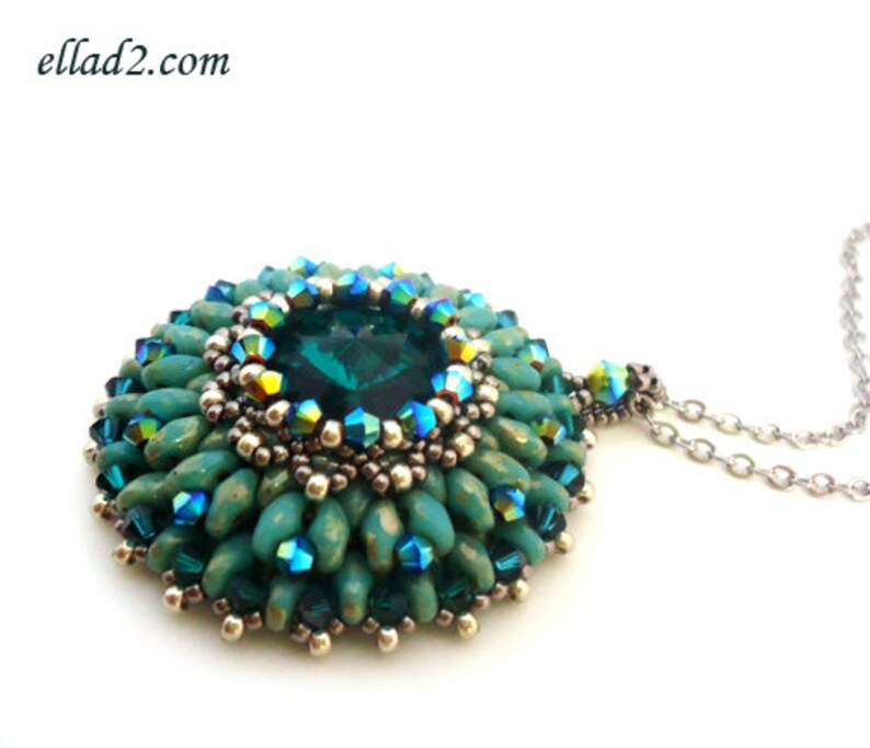 Op de afbeelding: Een ketting met een hanger van turquoise en groene kralen, met in het midden een grote, gefacetteerde, donkergroene edelsteen. De hanger is omringd door kleinere, iriserende kralen.