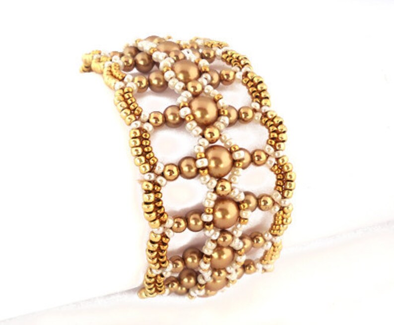 K&ouml;nnte beinhalten: Ein goldfarbenes und wei&szlig;es Armband mit geometrischem Muster. Das Armband besteht aus kleinen runden Perlen und gr&ouml;&szlig;eren runden Perlen.