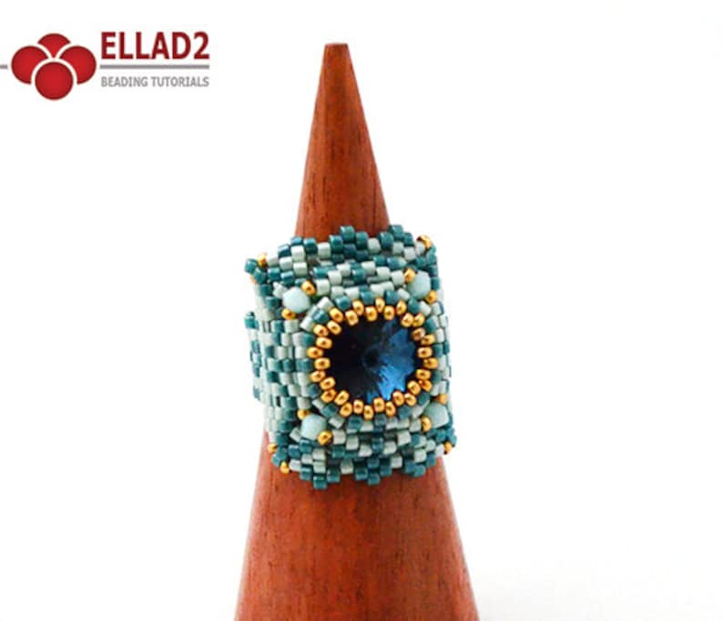 May include: A square-shaped beaded ring with a blue gemstone in the center, surrounded by gold beads. The ring is on a wooden ring stand.