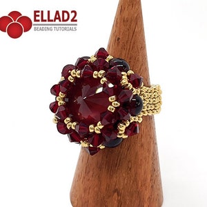 Może przedstawiać: Pierścionek z czerwonych i złotych koralik&oacute;w z dużym czerwonym kryształem w środku. Pierścionek znajduje się na drewnianym stojaku na pierścionki.