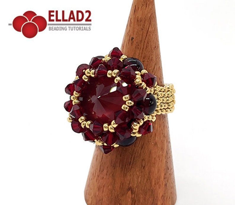 Może przedstawiać: Pierścionek z czerwonych i złotych koralik&oacute;w z dużym czerwonym kryształem w środku. Pierścionek znajduje się na drewnianym stojaku na pierścionki.