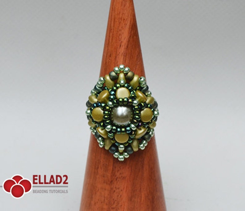 May include: A green and gold beaded ring with a pearl in the center, displayed on a wooden ring stand.