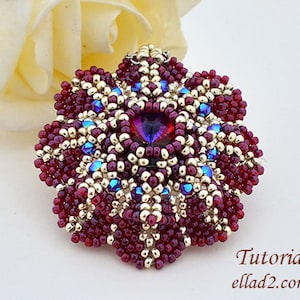 Pode incluir: Um pingente de flor de contas vermelhas, douradas e azuis com um grande cristal roxo iridescente no centro. O pingente é feito com contas de sementes e é um belo exemplo de tecelagem de contas.