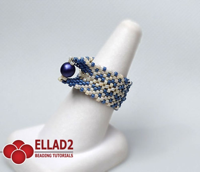 Peut inclure: Bague perl&eacute;e bleue et blanc cass&eacute;, orn&eacute;e d'une perle bleu fonc&eacute;. La bague est pr&eacute;sent&eacute;e sur un support blanc en forme de doigt. La bague est faite de petites perles carr&eacute;es tiss&eacute;es ensemble selon un motif g&eacute;om&eacute;trique. Le logo "ELLAD2 BEADING TUTORIALS" est en bas &agrave; gauche.