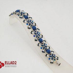 Op de afbeelding: Een blauwe en zilveren kralenarmband met een bloemenpatroon. De armband ligt op een witte displaystandaard. De armband is gemaakt van kleine, ronde kralen en grotere, ovale kralen. De kralen zijn gerangschikt in een patroon dat lijkt op bloemen.