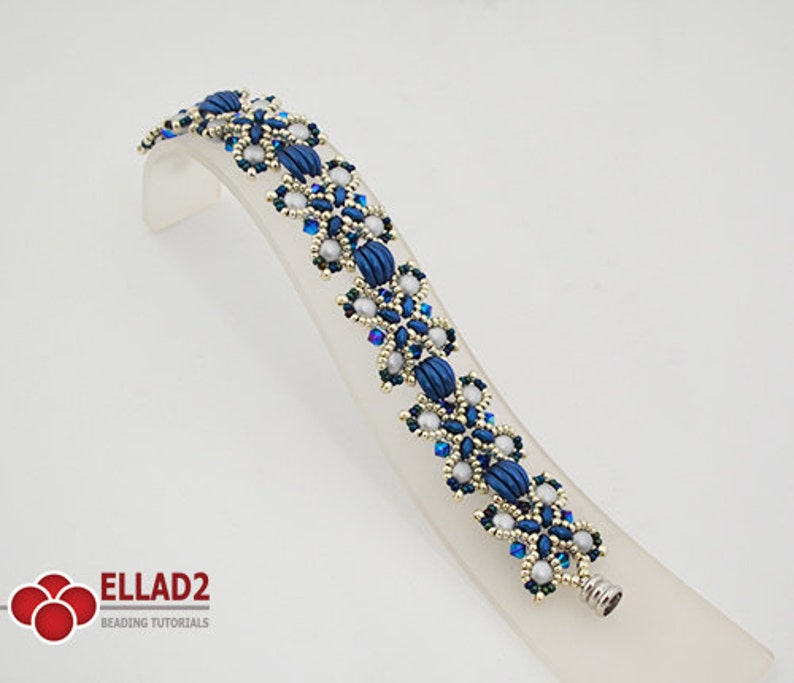 Op de afbeelding: Een blauwe en zilveren kralenarmband met een bloemenpatroon. De armband ligt op een witte displaystandaard. De armband is gemaakt van kleine, ronde kralen en grotere, ovale kralen. De kralen zijn gerangschikt in een patroon dat lijkt op bloemen.