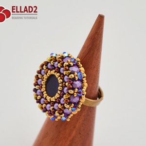 K&ouml;nnte beinhalten: Ein lila-goldener Perlenring mit schwarzem Zentrum. Der Ring befindet sich auf einem h&ouml;lzernen Ringst&auml;nder.