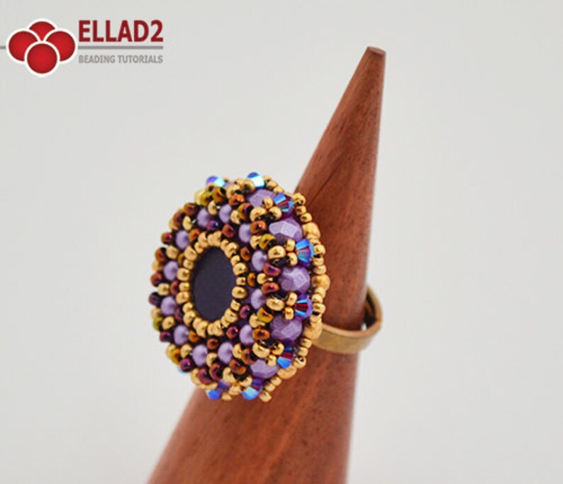 K&ouml;nnte beinhalten: Ein lila-goldener Perlenring mit schwarzem Zentrum. Der Ring befindet sich auf einem h&ouml;lzernen Ringst&auml;nder.
