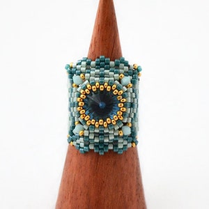 May include: A teal and light blue beaded ring with a gold-rimmed blue crystal in the center. The ring is displayed on a wooden ring stand.
