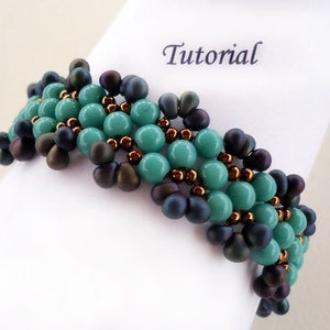 May include: A turquoise and dark blue beaded bracelet with a gold accent. The bracelet is on a white surface with the word "Tutorial" written on it.