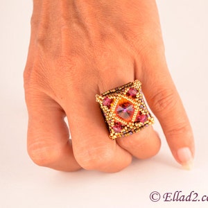 May include: A square-shaped ring with a large, faceted, orange gemstone surrounded by smaller, pink gemstones. The ring is made of gold-colored beads and is worn on a person's finger.
