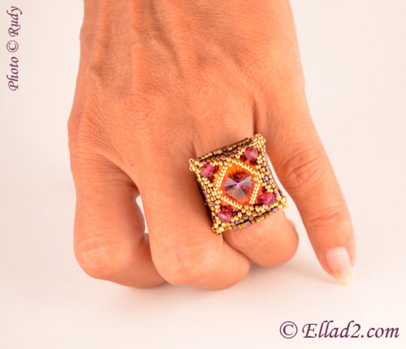 May include: A square-shaped ring with a large, faceted, orange gemstone surrounded by smaller, pink gemstones. The ring is made of gold-colored beads and is worn on a person's finger.