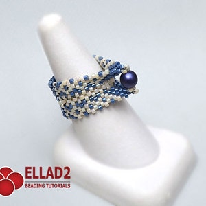 Peut inclure: Bague perl&eacute;e faite &agrave; la main, avec un motif tiss&eacute; de perles bleues et blanc cass&eacute;. Une perle bleu fonc&eacute; orne le dessus. La bague est pr&eacute;sent&eacute;e sur un support blanc en forme de doigt. Le logo "ELLAD2 BEADING TUTORIALS" est visible en bas &agrave; gauche.