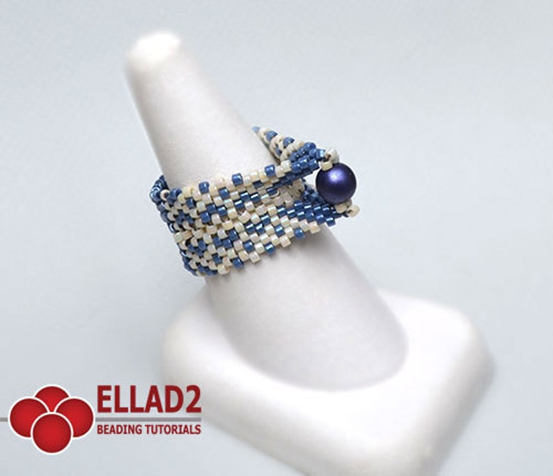 Peut inclure: Bague perl&eacute;e faite &agrave; la main, avec un motif tiss&eacute; de perles bleues et blanc cass&eacute;. Une perle bleu fonc&eacute; orne le dessus. La bague est pr&eacute;sent&eacute;e sur un support blanc en forme de doigt. Le logo "ELLAD2 BEADING TUTORIALS" est visible en bas &agrave; gauche.