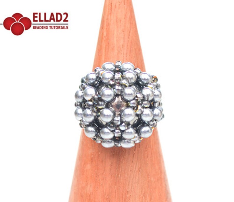 May include: A silver beaded ring with a round, spherical design. The ring is displayed on a wooden ring stand.