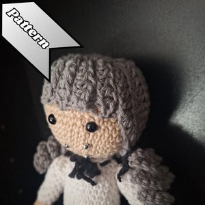 Könnte beinhalten: Gehäkelte Puppe mit grauem Haar, einem beigen Gesicht und schwarzen Augen. Die Puppe trägt ein weißes Hemd, einen weinroten Rock und eine schwarze Schleife. Das Wort "Pattern" befindet sich in einem weißen Banner oben links.