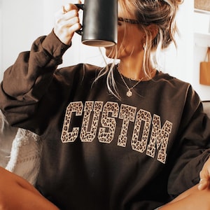 May include: A dark chocolate brown crewneck sweatshirt featuring the word CUSTOM in a block font with a leopard print pattern across the chest. The garment has long sleeves and a ribbed neckline. A small circular graphic in the corner reads Dark Chocolate.