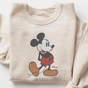May include: Sand-colored sweatshirt featuring a classic Mickey Mouse graphic. The cartoon character is depicted in black, red, and brown, with the Disney logo. The sweatshirt has a crew neck and long sleeves.