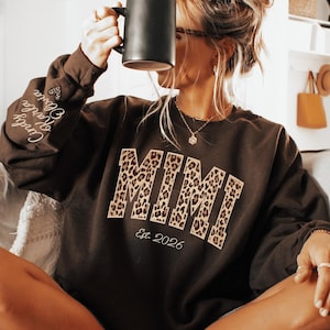 May include: Dark chocolate-colored sweatshirt with the word "MIMI" in leopard print and "Est. 2026" below. The sleeve has embroidered names. The sweatshirt is worn by a person holding a black mug.