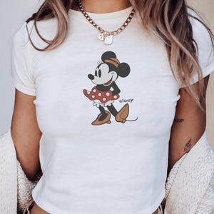 Pu&ograve; includere: Top corto bianco con grafica di Minnie Mouse. Il personaggio dei cartoni animati &egrave; raffigurato in una posa classica, con una gonna a pois rossi e scarpe marroni. Il logo Disney &egrave; stampato sotto.