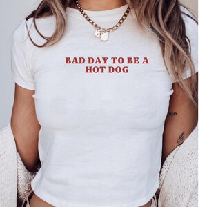 Puede incluir: Top corto blanco con la frase "BAD DAY TO BE A HOT DOG" en rojo. La camiseta tiene mangas cortas y cuello redondo. La persona lleva un collar dorado y una rebeca color crema.