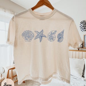 May include: Ivory-colored t-shirt featuring blue seashell and starfish designs. The short-sleeved shirt is hanging on a wooden hanger. The shirt's design includes a seashell, starfish, and two other shell illustrations.