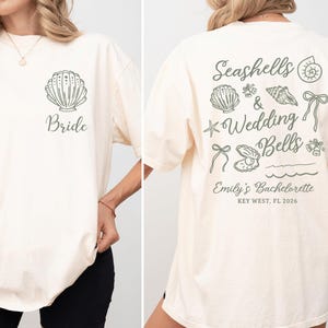 May include: Ivory-colored t-shirt with a seashell graphic and the word "Bride" on the front. The back features "Seashells & Wedding Bells" text and seashell illustrations. "Emily's Bachelorette, Key West, FL 2026" is also printed on the back.