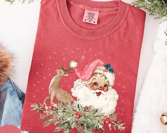 May include: A red t-shirt featuring a vintage Santa Claus and reindeer design. The graphic includes a smiling Santa with a pink hat and a reindeer standing on a bed of pine needles and berries. The shirt is surrounded by Christmas decorations.
