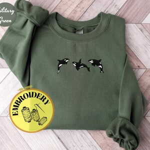 May include: A military green sweatshirt with embroidered orca whales. The sweatshirt features three black and white orcas in a row. The words "Military Green" are in a circle on the top left. The word "EMBROIDERY" is in a circle on the bottom left.
