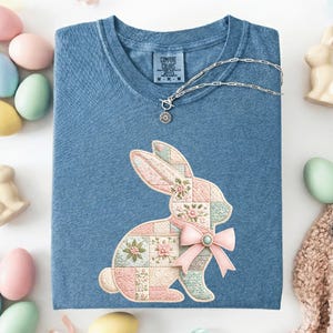 May include: A blue Comfort Colors t-shirt with a patchwork bunny design and a pink bow. A silver necklace is draped on the shirt. Pastel Easter eggs and ceramic bunnies create a spring theme. The shirt is a comfortable fit.