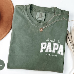 May include: Moss green t-shirt with the text "Amelia's PAPA EST. 2026" in white block letters. The shirt has a crew neck and short sleeves. A gold necklace with a pendant is draped on the shirt.