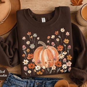 May include: A dark chocolate brown crewneck sweatshirt featuring a central graphic of a large orange pumpkin surrounded by a variety of small, colourful wildflowers in shades of orange, white, and pink. The garment has long sleeves and a ribbed collar.