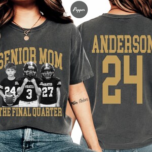 May include: A charcoal gray t-shirt featuring a graphic of three children in football uniforms with the text SENIOR MOM THE FINAL QUARTER on the front, and the name ANDERSON with the number 24 printed in gold on the back of the garment.