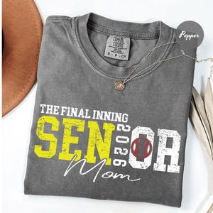 May include: A gray Comfort Colors t-shirt with the text "THE FINAL INNING SENIOR 2026 Mom" in yellow and white. A baseball graphic is incorporated into the "O" of "Mom". A silver necklace with a pendant is on the shirt.