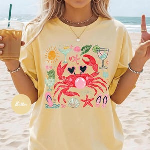 May include: Yellow t-shirt featuring a vibrant graphic of a red crab with hearts, cocktails, seashells, and a sun. The design evokes a beach or summer theme, perfect for casual wear.