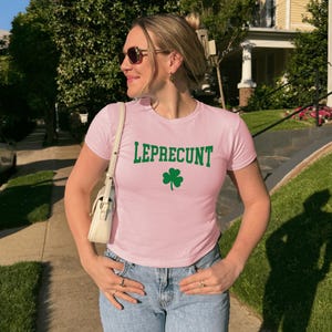 May include: A pale pink t-shirt featuring the word "LEPRECUNT" in green lettering and a green shamrock. The shirt is paired with blue denim jeans.