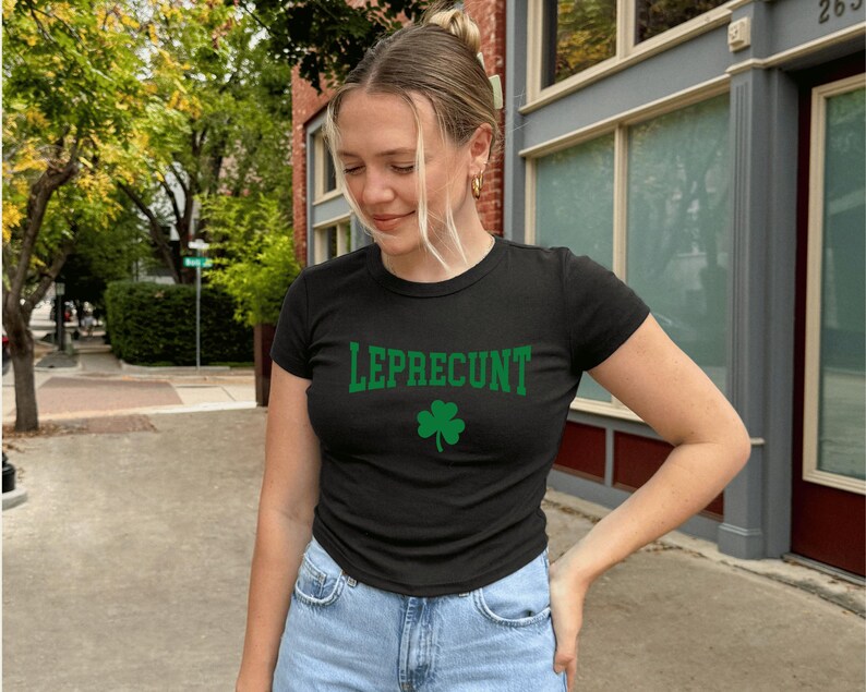 May include: Black crew neck t-shirt with the word "LEPRECONT" in green block letters and a green shamrock. The shirt is worn by a person in blue jeans, standing outside on a sunny day.
