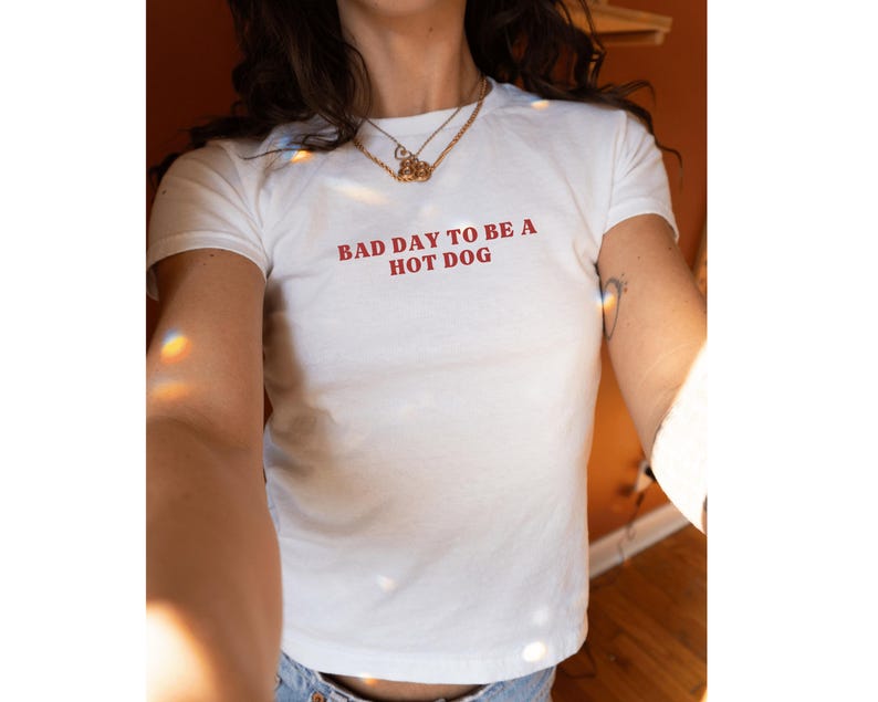Puede incluir: Camiseta blanca con la frase roja "BAD DAY TO BE A HOT DOG". La camiseta de manga corta tiene cuello redondo. La persona lleva un collar dorado. La camiseta es de material suave.