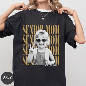 May include: A black t-shirt featuring a graphic print with the repeated gold text SENIOR MOM. Centered is a black and white photograph of a child wearing heart-shaped sunglasses and making a peace sign, with the name Amelia written in white script.
