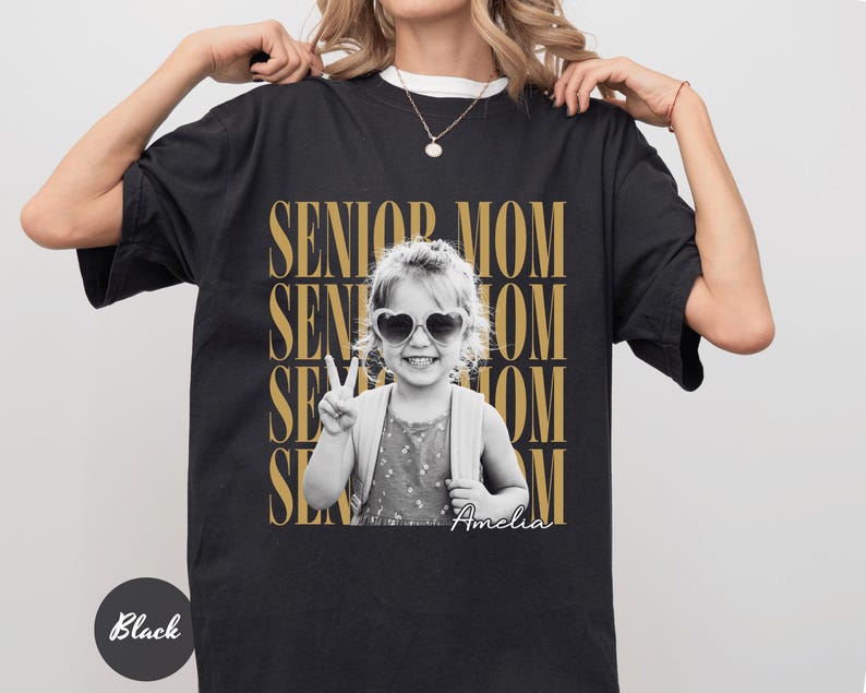 May include: A black t-shirt featuring a graphic print with the repeated gold text SENIOR MOM. Centered is a black and white photograph of a child wearing heart-shaped sunglasses and making a peace sign, with the name Amelia written in white script.