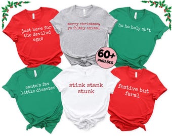 Op de afbeelding: Zes shirts met korte mouwen in rood, grijs, groen en wit. Elk shirt heeft een andere humoristische zin in witte of rode tekst, waaronder "just here for the deviled eggs" en "merry christmas, ya filthy animal".