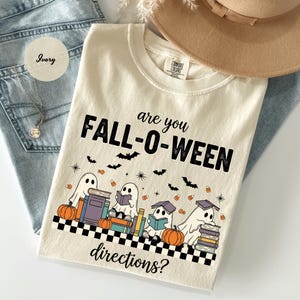 May include: Ivory-colored t-shirt with the text "are you FALL-O-WEEN directions?" featuring ghost illustrations, pumpkins, books, and bats. The shirt is laid on top of blue jeans and a tan hat.