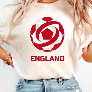 May include: Cream-colored t-shirt featuring a red and white soccer ball graphic with the word "ENGLAND" in red block letters below. The soccer ball design is a stylized representation of a soccer ball.