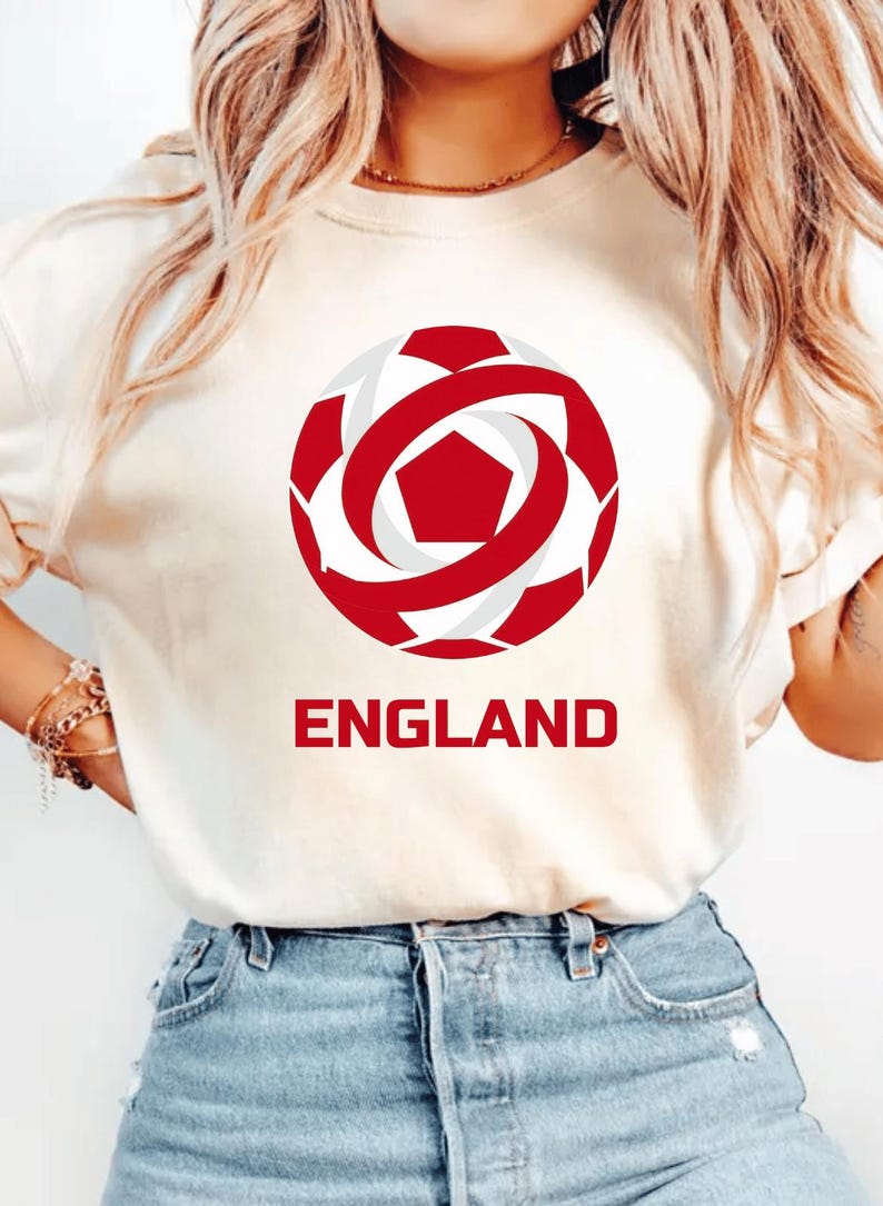 May include: Cream-colored t-shirt featuring a red and white soccer ball graphic with the word "ENGLAND" in red block letters below. The soccer ball design is a stylized representation of a soccer ball.