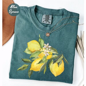 May include: A teal-colored t-shirt featuring a watercolor illustration of lemons with green leaves and white blossoms. The shirt has a crew neck and short sleeves. A gold necklace with a pendant rests on the shirt. The words "Blue Spruce" are in a circle on the top left.