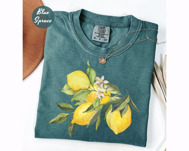 May include: A teal-colored t-shirt featuring a watercolor illustration of lemons with green leaves and white blossoms. The shirt has a crew neck and short sleeves. A gold necklace with a pendant rests on the shirt. The words "Blue Spruce" are in a circle on the top left.
