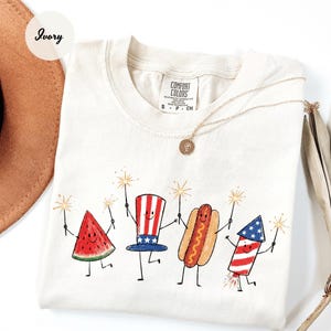 K&ouml;nnte beinhalten: Elfenbeinfarbenes T-Shirt mit einer skurrilen Illustration einer Wassermelonenscheibe, einem Uncle-Sam-Hut, einem Hotdog und einem Feuerwerk, die jeweils Wunderkerzen halten. Das Comfort Colors-Shirt hat einen Rundhalsausschnitt und kurze &Auml;rmel. Eine goldene Halskette ist &uuml;ber dem Shirt drapiert.
