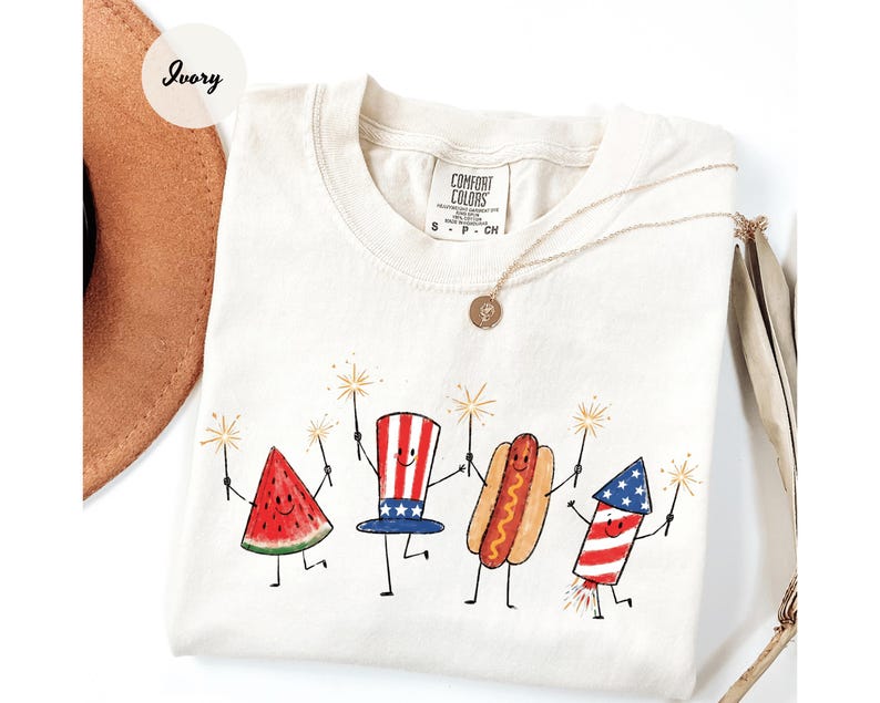 K&ouml;nnte beinhalten: Elfenbeinfarbenes T-Shirt mit einer skurrilen Illustration einer Wassermelonenscheibe, einem Uncle-Sam-Hut, einem Hotdog und einem Feuerwerk, die jeweils Wunderkerzen halten. Das Comfort Colors-Shirt hat einen Rundhalsausschnitt und kurze &Auml;rmel. Eine goldene Halskette ist &uuml;ber dem Shirt drapiert.