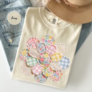 May include: Ivory-colored t-shirt with a floral applique design. The applique features various fabric patterns in pink, blue, yellow, and green. The shirt is laid on top of a pair of blue jeans, with a necklace and a tan hat in the background.