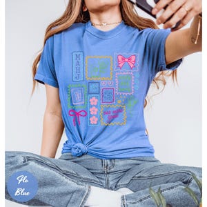 May include: A blue t-shirt with a graphic design featuring various postage stamp-like images. The stamps include a pink bow, flowers, and the text "one more game". The shirt is tied at the waist.